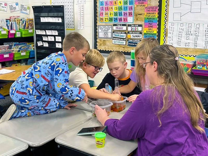 A teacher works with four elementary students gathered around a classroom table on a hands-on project.